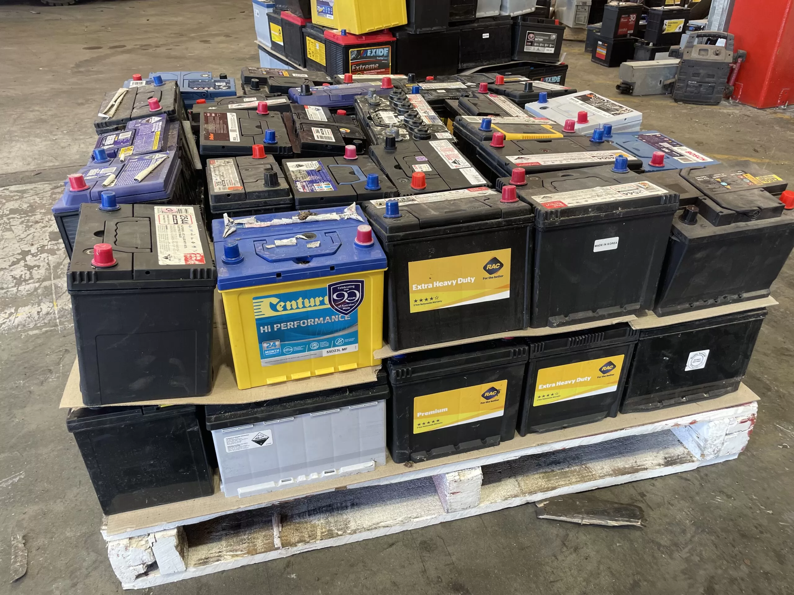 A pallet stacked with various car batteries of different brands, colors, and sizes sits inside a workshop—perfect for those looking to sell scrap or 回收. More batteries are visible in the background, emphasizing the range available.