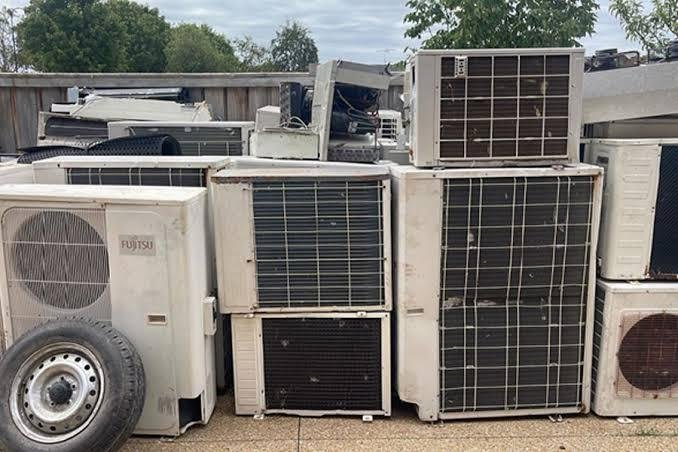 A stack of old, used air conditioning units and parts, including a spare tire, arranged outdoors on a paved surface with trees and a wooden fence in the background—perfect for those looking to Sell Scrap or 回收 valuable materials.